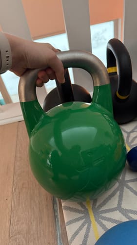 Customer holding a shiny green Competition Kettlebell by its steel handle indoors.