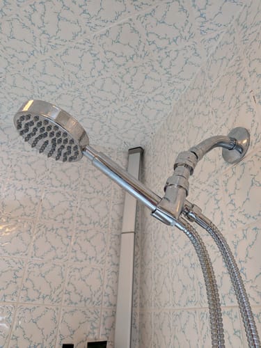 Reviewer's shiny Shower Head Swivel Adapter installed on a shower head, allowing for a greater downward angle.