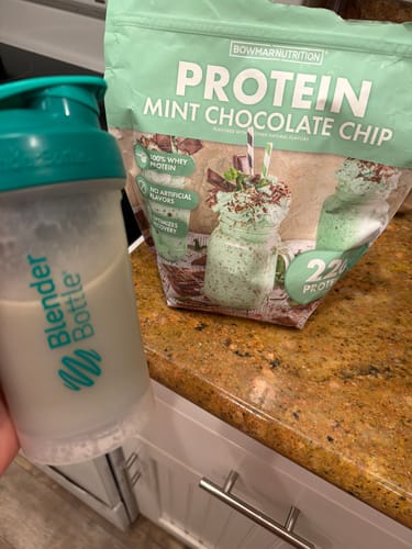 Customer holding a shaker cup with a mixed shake next to a bag of Mint Chocolate Chip Whey Protein on a counter.