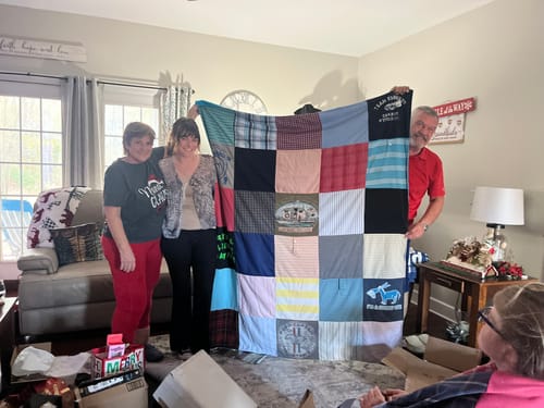 Customers holding up a large, colorful t-shirt quilt made from many different shirts in a living room.