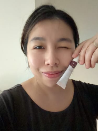 Customer winks while holding a Super Peptide SPF Lip Tint, showing the product's glossy, tinted finish on her lips.