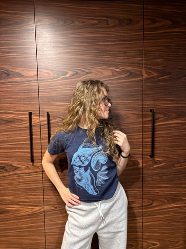 Reviewer wearing a dark sandy baby tee with a blue graphic, standing indoors in front of a wooden wall.