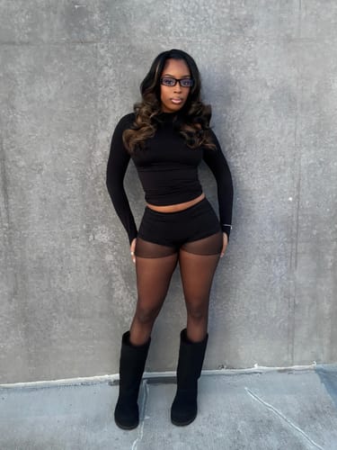 Customer wearing the black Seamless Micro Mini Shorts with a matching top, sheer tights, and boots against a grey wall.
