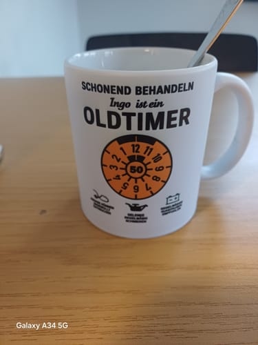 Rezensent zeigt die weiße, personalisierte OLDTIMER-Tasse mit dem Namen 'Ingo' und einem TÜV-Plaketten-Motiv auf einem Tisch.