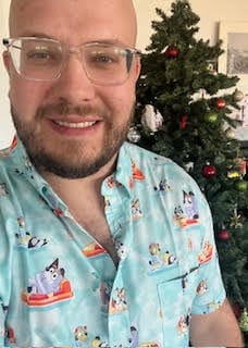 Customer wearing the light blue Bluey short sleeve shirt in front of a decorated Christmas tree.
