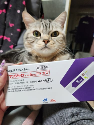 レビュアーが手に持っている「マンジャロ皮下注5mgアテオス」の箱と、その後ろに座っている猫。