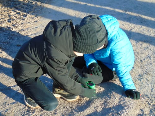 Zwei Kinder eines Kunden untersuchen draußen auf einem Weg etwas mit dem grünen Edubini Abenteuer-Mikroskop.