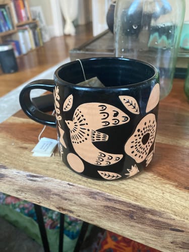 Customer's black Myth Imprint Stoneware Mug with a teabag inside, sitting on a wooden table.