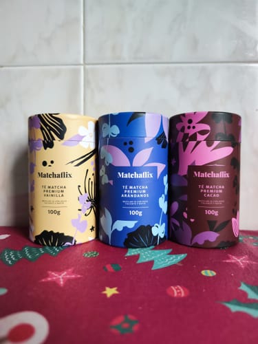El cliente muestra el Té Matcha Vainilla Premium junto con los de arándanos y cacao sobre una superficie roja.