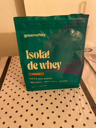 Un client montre le sachet vert d'Isolat de Whey Native saveur chocolat, posé sur une table à pois.