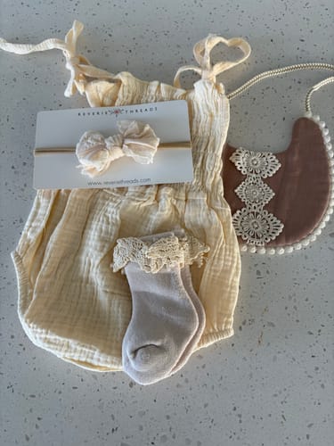 Customer's cream-colored baby outfit displayed flat, including a romper, headband, lace socks, and decorative bib.