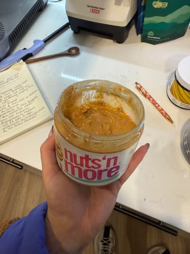 Reviewer holding a partially used jar of Birthday Cake High Protein Peanut Butter Spread in a kitchen.