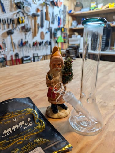 Customer's clear glass beaker bong with a teal mouthpiece sitting on a wooden workbench.