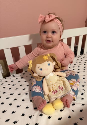 A reviewer's baby sits in a crib holding the Personalized Daisy Girl doll, showing the name embroidered on its yellow dress.