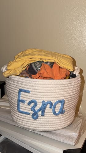 Customer's personalized knitted basket in white, with the name 'Ezra' in blue yarn and filled with clothing.