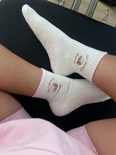 A reviewer wearing the white 'All I Want Is Pilates' socks with pink flower-shaped grips on a Pilates reformer.