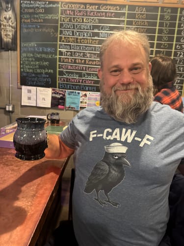 Reviewer smiling at a bar while wearing the blue F-CAW-F t-shirt with a crow in a sailor hat graphic.