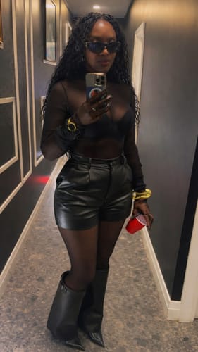 Reviewer posing in black Pointed Toe Vegan Leather Knee Boots, styled with leather shorts and a sheer top.