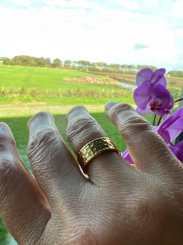 Customer's hammered gold Amaya ring worn on a finger against a blurred outdoor landscape with purple flowers