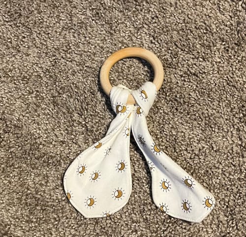 Reviewer's teething ears sun ray teether on carpeted floor, showing a simple wooden ring with cotton fabric ears.