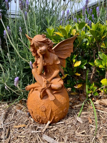 Customer's rust-colored Lady Ophelia fairy statue sitting on a flowered sphere in a garden with lavender.