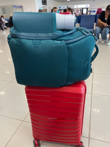Customer's teal The Underseat 15L bag sitting on top of a red suitcase in an airport terminal.