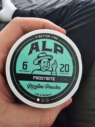 Customer's hand holding a round tin of ALP Frostbite nicotine pouches with teal and black branding.