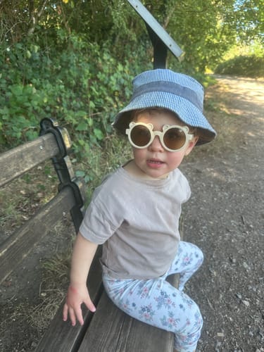 Customer's child wearing sunglasses and a patterned bucket hat, sitting on a wooden bench outdoors.