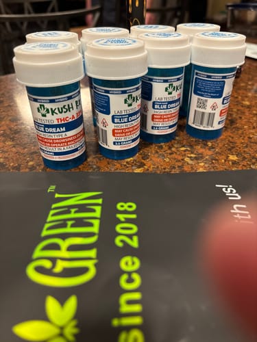 Customer's several Blue Dream Kush Rx flower bottles on a brown surface with a partial blurred brand flyer in front.