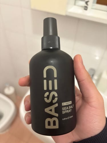 Customer's hand holding a black, cylindrical BASED Sea Salt Spray bottle in a bathroom setting.