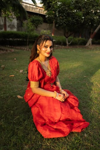 Customer's Gouri’s Grace Jamdani Saree in vibrant red, worn by a person sitting on a grassy lawn.