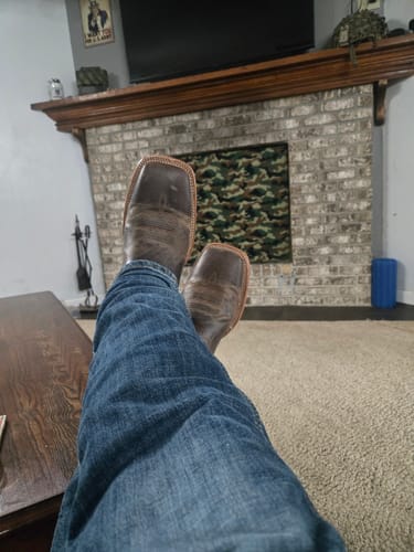 Customer's brown wide square toe western boots worn by a person resting feet on a fireplace hearth.
