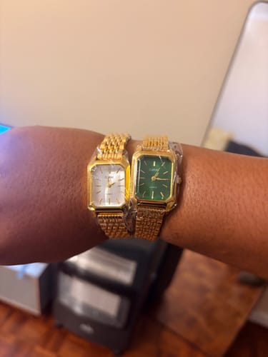 Customer's hand wearing two Claire De Mains gold-tone watches with rectangular faces in silver and green.
