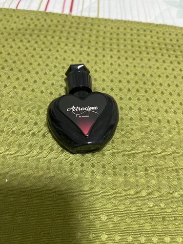 Customer's Attracione Sensual perfume in a black, heart-shaped bottle, sitting on a textured green surface.