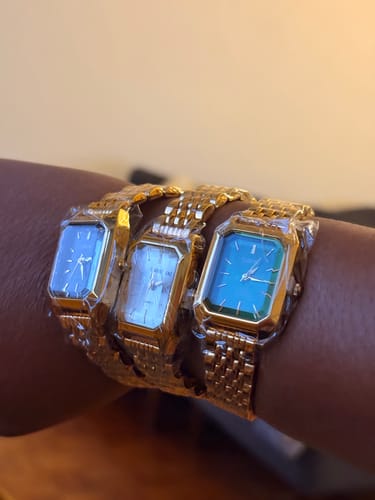 Customer's hand wearing three Claire De Mains gold-tone watches with rectangular faces in blue, white, and green.