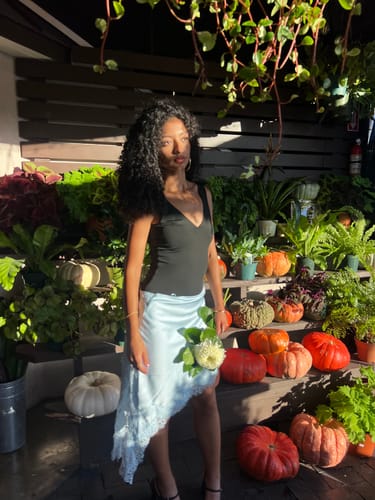 Reviewer wearing a black Jade Double Plunge Bodysuit with a light blue skirt while posing outdoors near pumpkins.