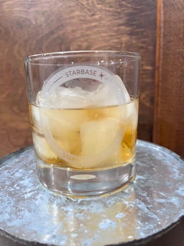 Customer's SpaceX Starship Flight 14 Whiskey Glass filled with amber liquid and ice cubes on a textured, metallic base.