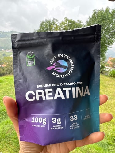 Customer's hand holding a blue and black package of Creatina Monohidrato outdoors with greenery in the background.