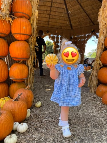 Customer's child wearing the blue gingham smocked bloomer set, holding a small pumpkin in a pumpkin patch.