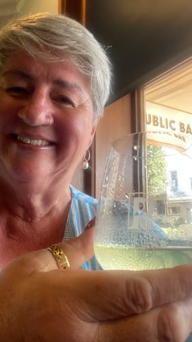 Reviewer holding a glass of clear Zero Sugar Chardonnay in a cafe setting.