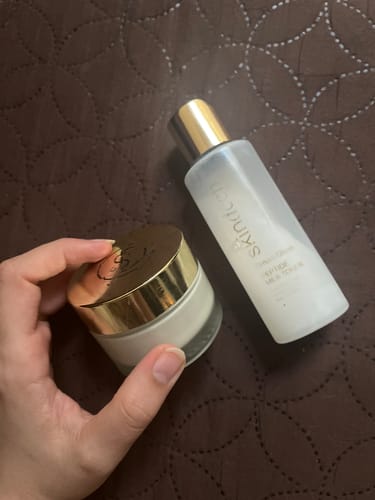 Reviewer's hand holding a DREAM GLAZE Peptide Milk Toner bottle and a small cream jar against a dark patterned background.