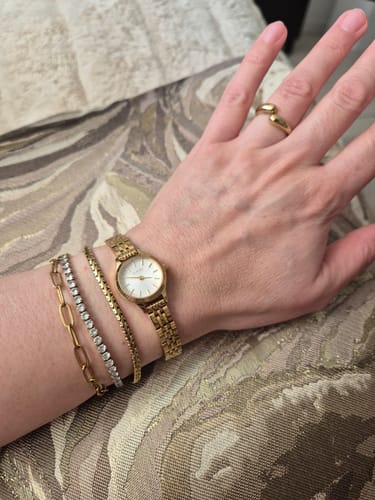 Customer's gold Sophie watch worn on a wrist alongside several other gold-toned bracelets and a gold ring.