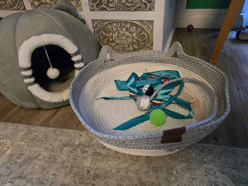 Reviewer's Fellnest Katzenkorb mit Spielzeug darin neben einer grauen Katzenhöhle auf einem Holzboden.