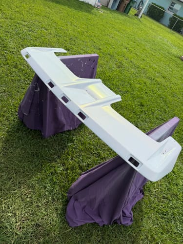 Customer's white painted car spoiler resting on a purple stand outdoors in a grassy yard.
