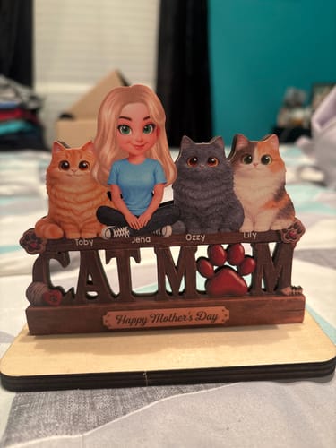 Customer's Cat Mom wooden plaque featuring a cartoon figure and three cats, customized with names, standing on a bed.
