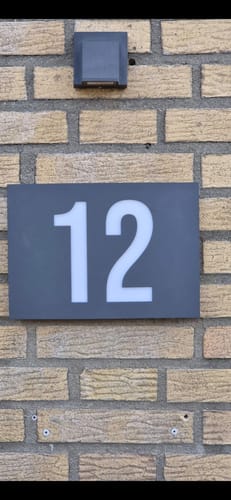Customer's montiertes, dunkles Hausnummernschild mit der Zahl 12 an einer hellen Backsteinwand.