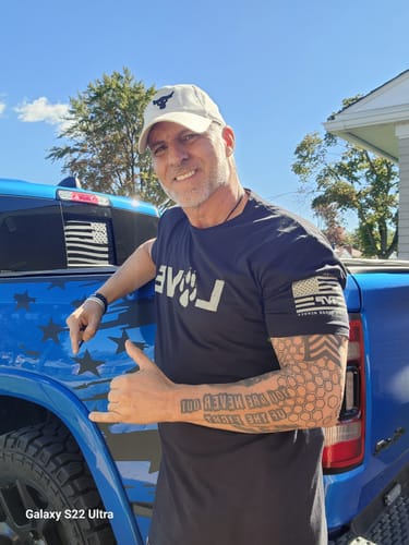 Customer's black The Love Of Dogs tee, featuring a white graphic, worn by a smiling person next to a blue truck.