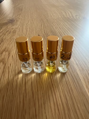 Customer's Tester Set of four small fragrance spray bottles with gold caps on a wooden table, some labels blurred.