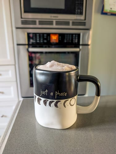 Customer's Chocolate Chili Chai tea latte served in a black and white moon-themed mug on a kitchen counter.