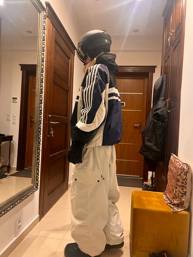 Reviewer's beige baggy waterproof snow pants worn with a jacket and helmet in a hallway mirror reflection